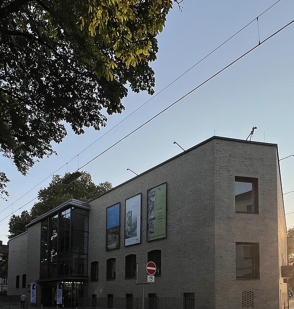 Blick auf ein Gebäude, im Vordergrund steht ein Baum (Anschnitt). Im Hintergrund ist ein Sonnenuntergang.