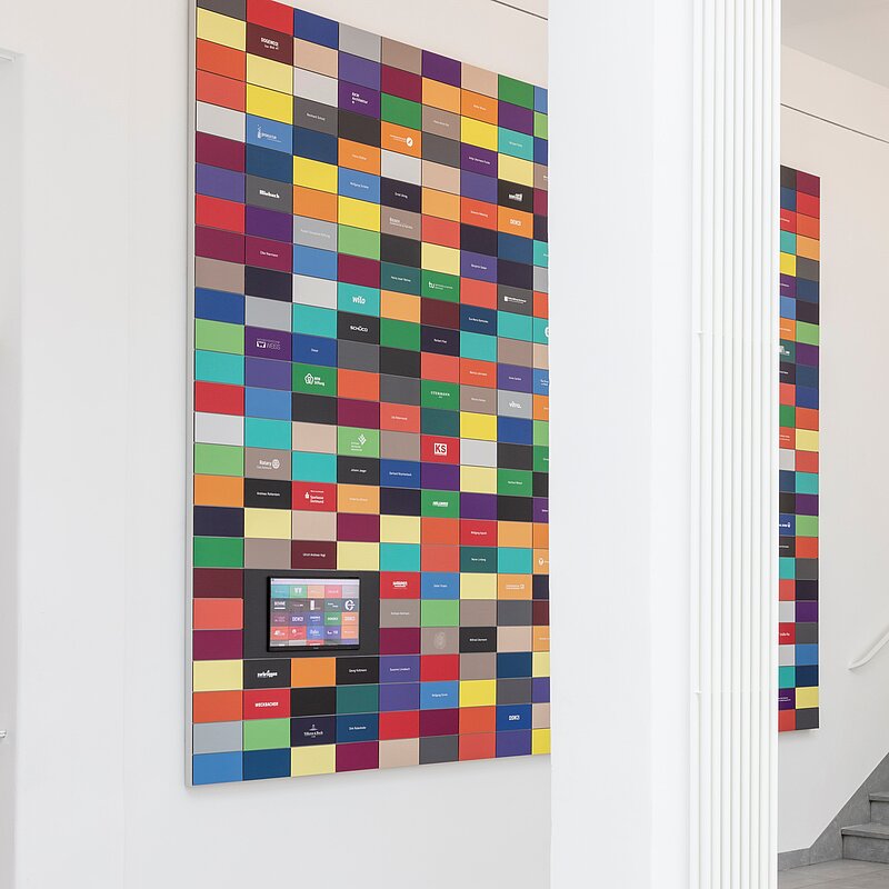 An einer hellen Wand hängen bunte Plaketten, auf denen Namen und Unternehmen abzulesen sind.
