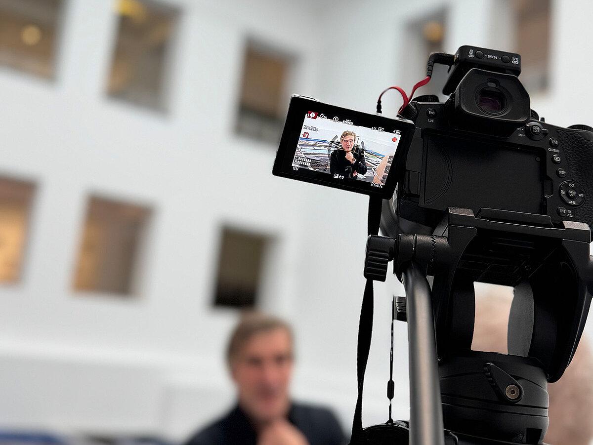 Zwei Personen stehen vor einer Kamera und führen ein Interview in einem hellen Raum.