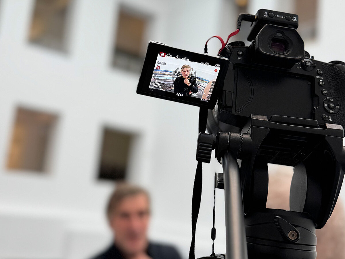 Zwei Personen stehen vor einer Kamera und führen ein Interview in einem hellen Raum.