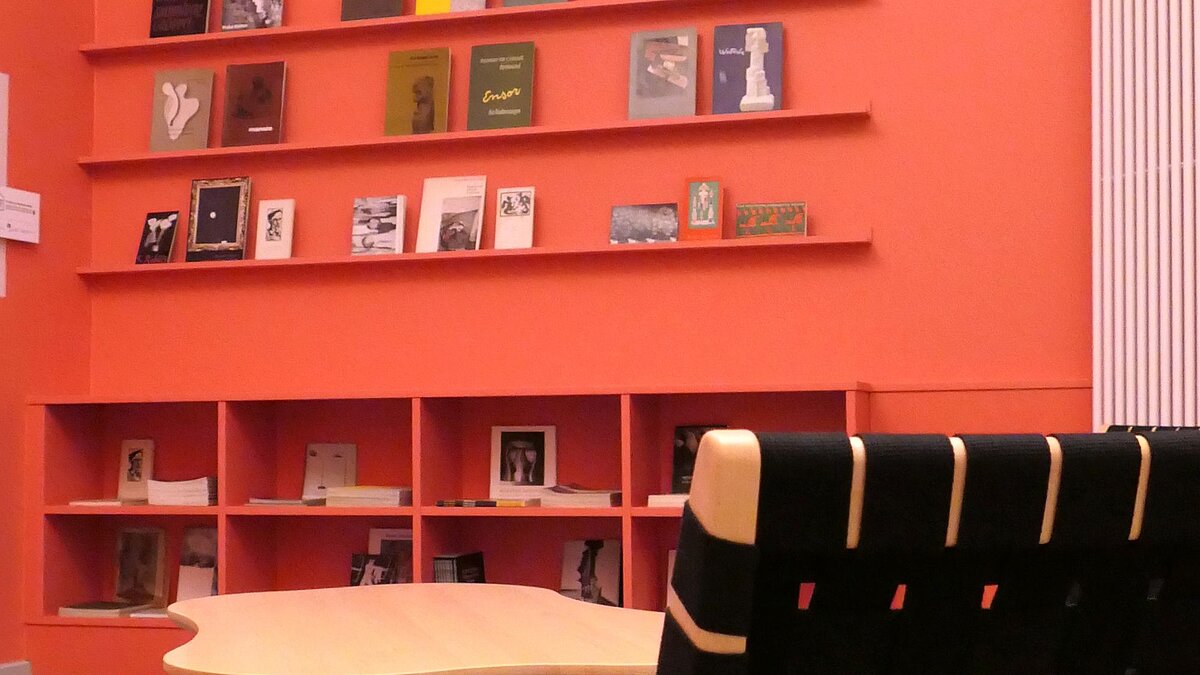 Ein Raum mit rötlichen Wänden, Bücherregalen, die mit Publikationen befüllt sind, einem Stuhl und einem Tisch.