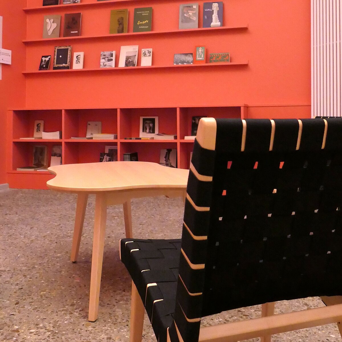 Ein Raum mit rötlichen Wänden, Bücherregalen, die mit Publikationen befüllt sind, einem Stuhl und einem Tisch.