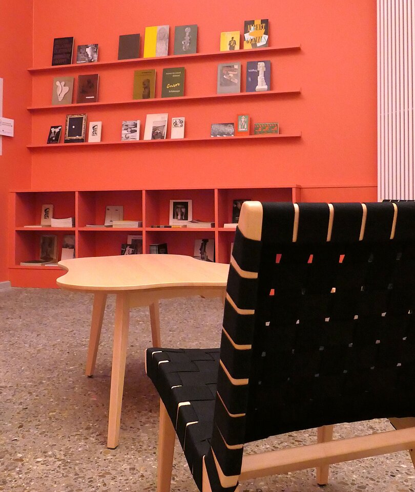 Ein Raum mit rötlichen Wänden, Bücherregalen, die mit Publikationen befüllt sind, einem Stuhl und einem Tisch.
