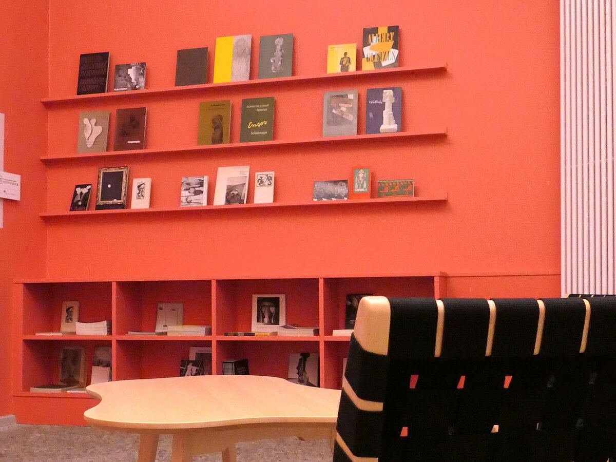 Ein Raum mit rötlichen Wänden, Bücherregalen, die mit Publikationen befüllt sind, einem Stuhl und einem Tisch.