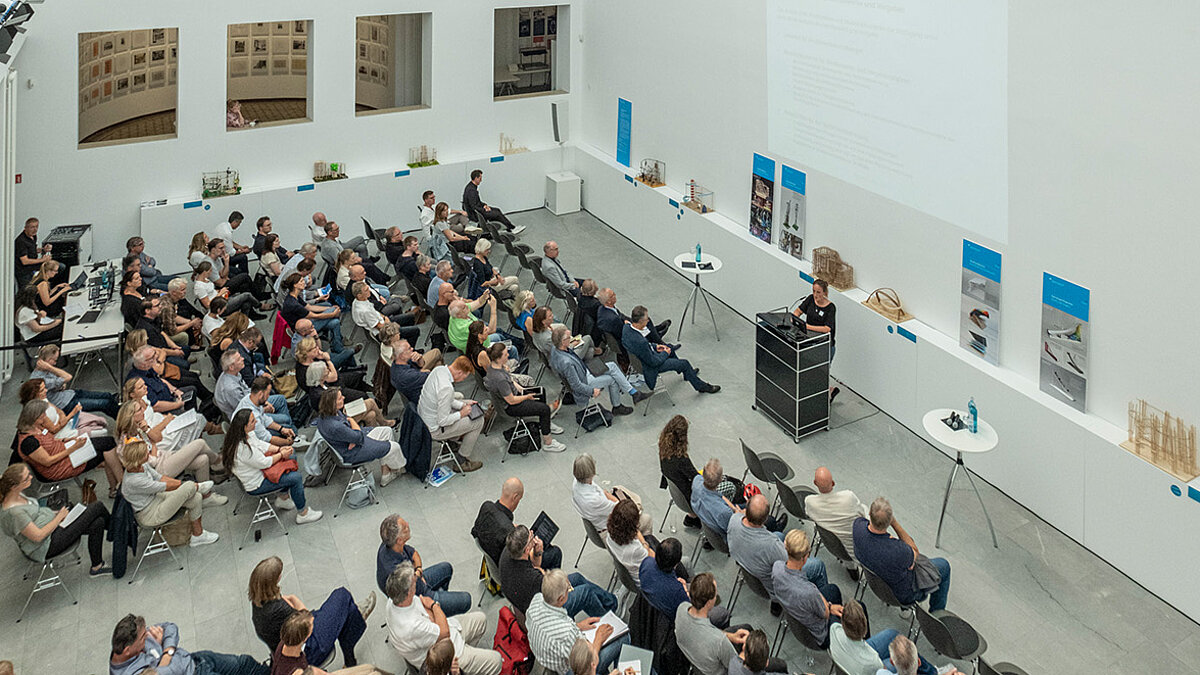 Ein heller Raum, viele Leute sitzen auf Stühlen im Publikum. An einem Pult steht eine Person und spricht zu dem Publikum. - Foto: Detlef Podehl