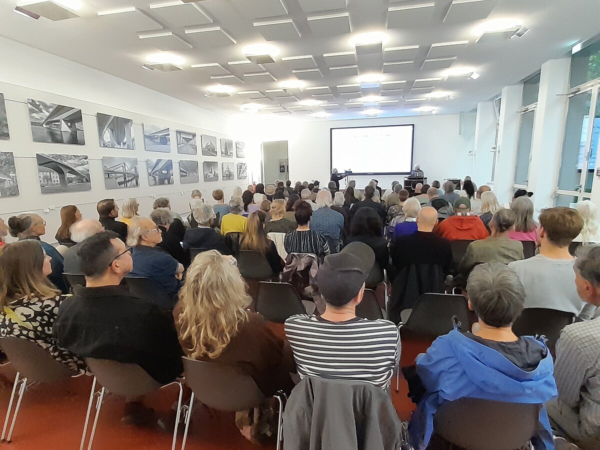 Ausstellungsraum mit Besuchenden, viele Leute sitzen auf Stühlen.