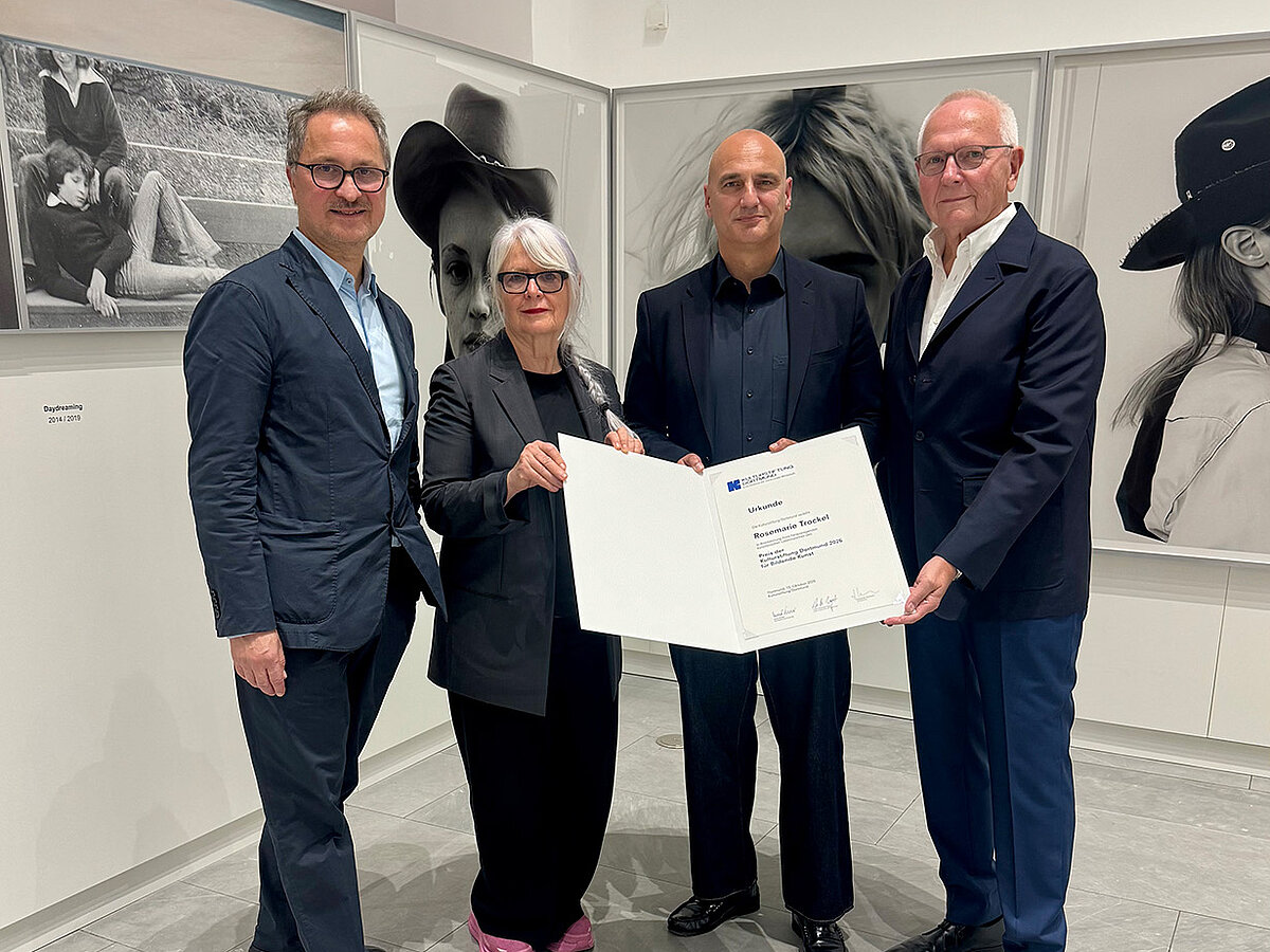Vier Personen in einem Ausstellungsraum. Die Personen stehen beisammen und einige von ihnen halten eine Urkunde fest und präsentieren diese in die Kamera. Im Hintergrund sind Kunstwerke an der Wand. 