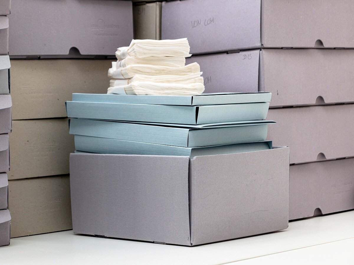 Graue sowie hellblaue Boxen und helle Dokumente liegen gestapelt nebeneinander und teilweise übereinander. -  Foto: Christine Kämmerer/Baukunstarchiv NRW