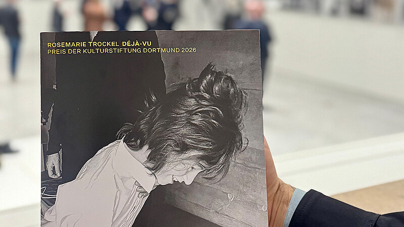 Heller Ausstellungsraum mit Publikum. Im Vordergrund wird eine Publikation gehalten.