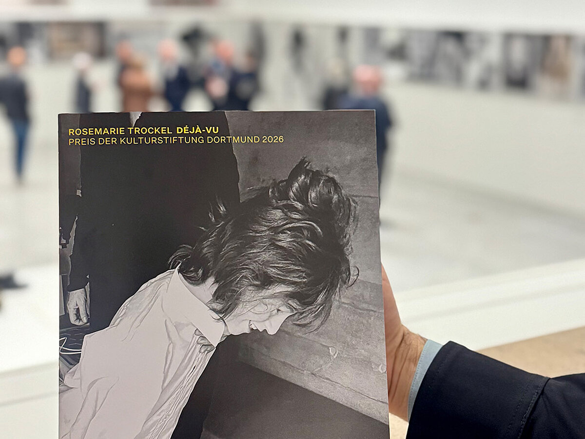 Heller Ausstellungsraum mit Publikum. Im Vordergrund wird eine Publikation gehalten.