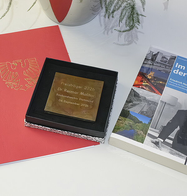 Auf einem Tisch liegen: Eine Urkunde, ein Buch und eine Plakette. Im Anschnitt ist ein Blumenstrauß.