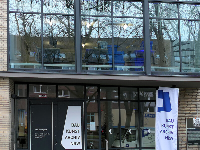 Blick auf die Eingangstür eines Gebäudes, oberhalb der Türe ist eine Glasfassade, vor der Tür steht eine Fahne. 