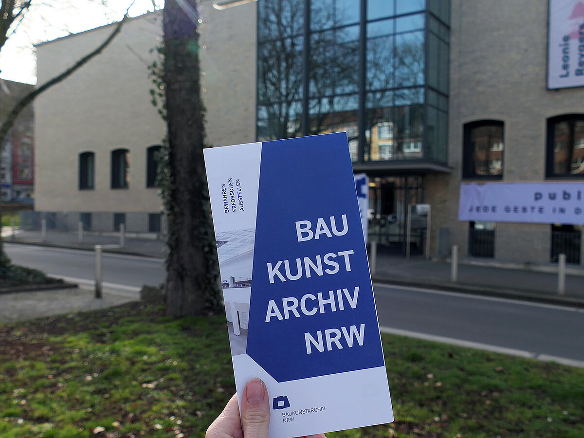 Ein Flyer wird von einer Hand hochgehalten, im Hintergrund ist ein großes Gebäude mit heller Fassade.