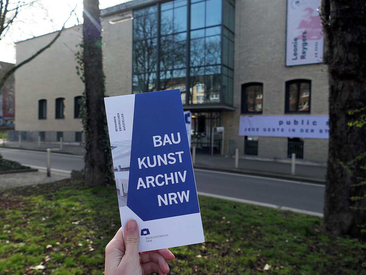 Ein Flyer wird von einer Hand hochgehalten, im Hintergrund ist ein großes Gebäude mit heller Fassade.