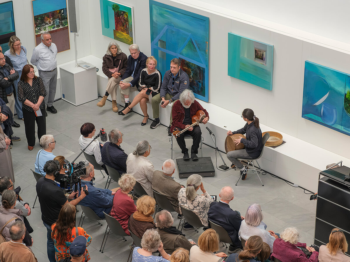 Personen sitzen und stehen in einem Ausstellungsraum, bunte Gemälde hängen an den Wänden.