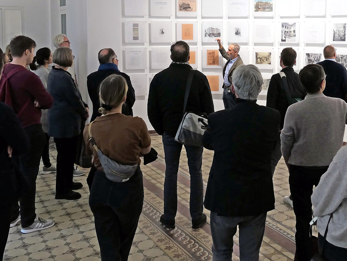 Eine Gruppe Personen steht in einem Raum, an der Wand hängen Fotos und Dokumente. Ein Mann spricht zu der Gruppe.
