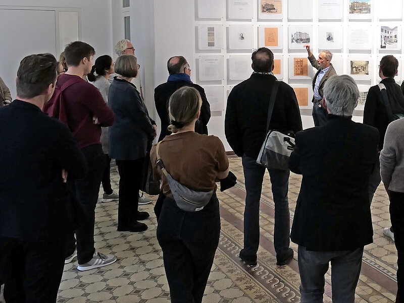 Eine Gruppe Personen steht in einem Raum, an der Wand hängen Fotos und Dokumente. Ein Mann spricht zu der Gruppe.