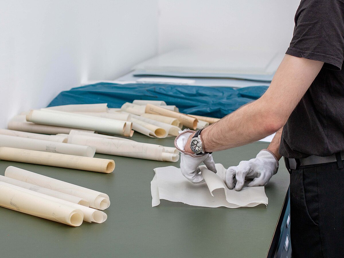 Baupläne aus Papier liegen aufgerollt nebeneinander, eine Person ist im Anschnitt zu sehen und arbeitet mit den Dokumenten.