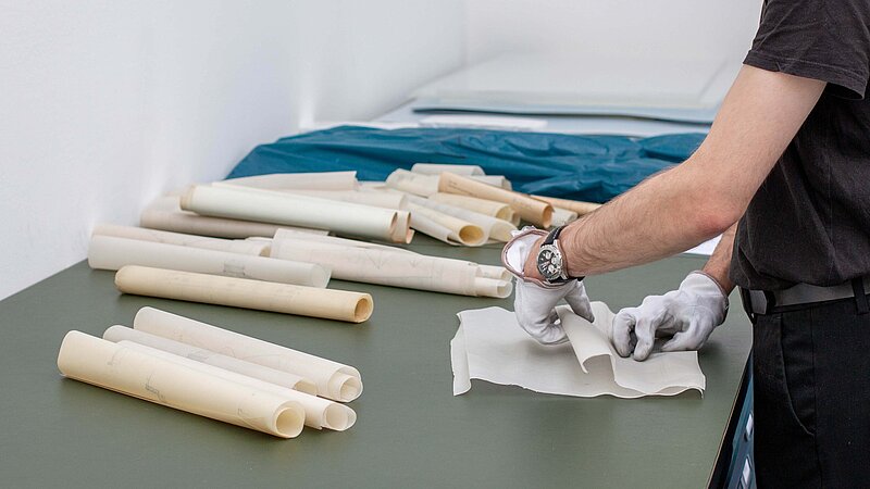 Baupläne aus Papier liegen aufgerollt nebeneinander, eine Person ist im Anschnitt zu sehen und arbeitet mit den Dokumenten.