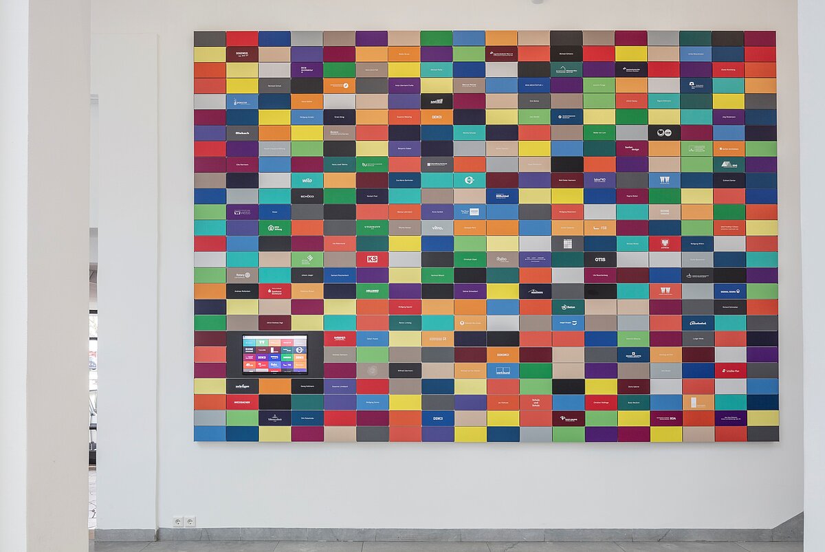An einer hellen Wand hängen bunte Plaketten, auf denen Namen und Unternehmen abzulesen sind.