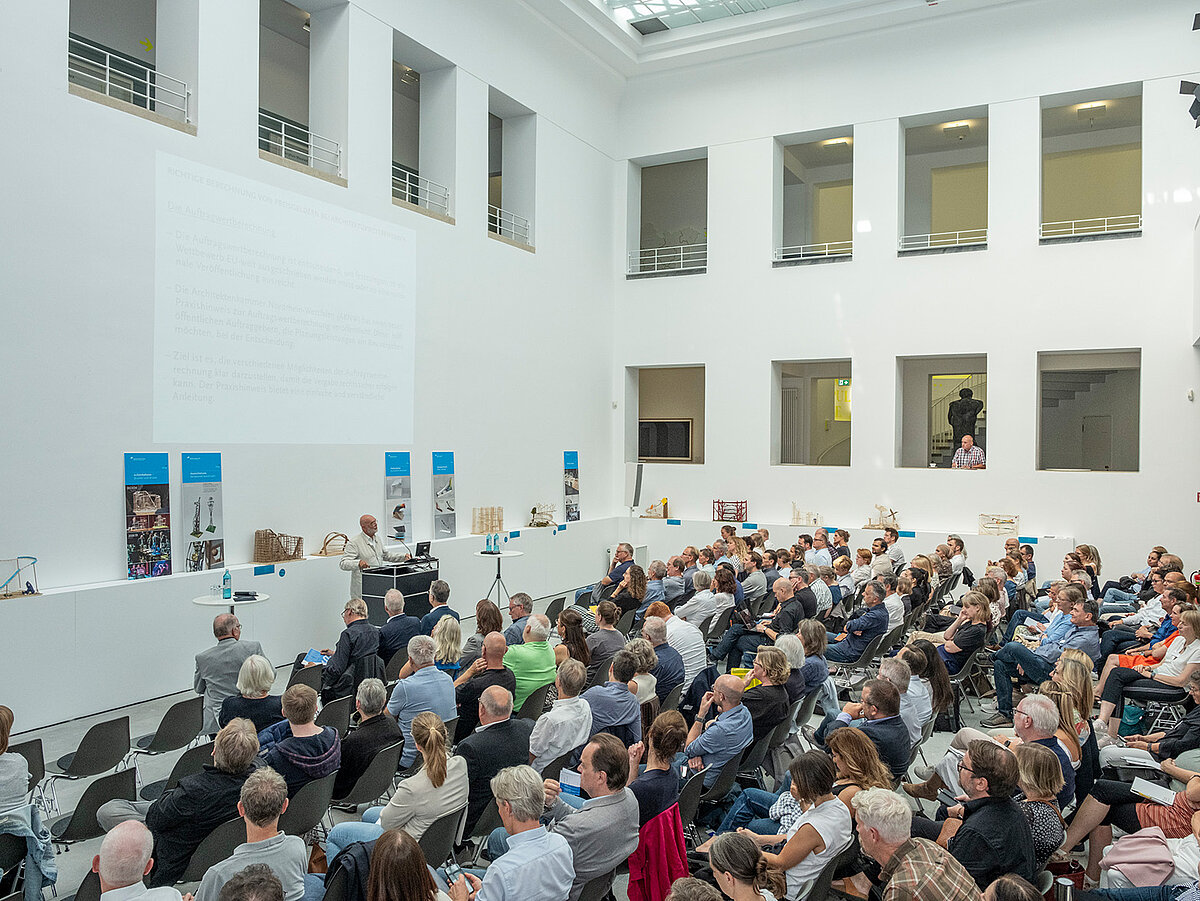 Ein heller Raum, viele Leute sitzen auf Stühlen im Publikum. An einem Pult steht eine Person und spricht zu dem Publikum. - Foto: Detlef Podehl