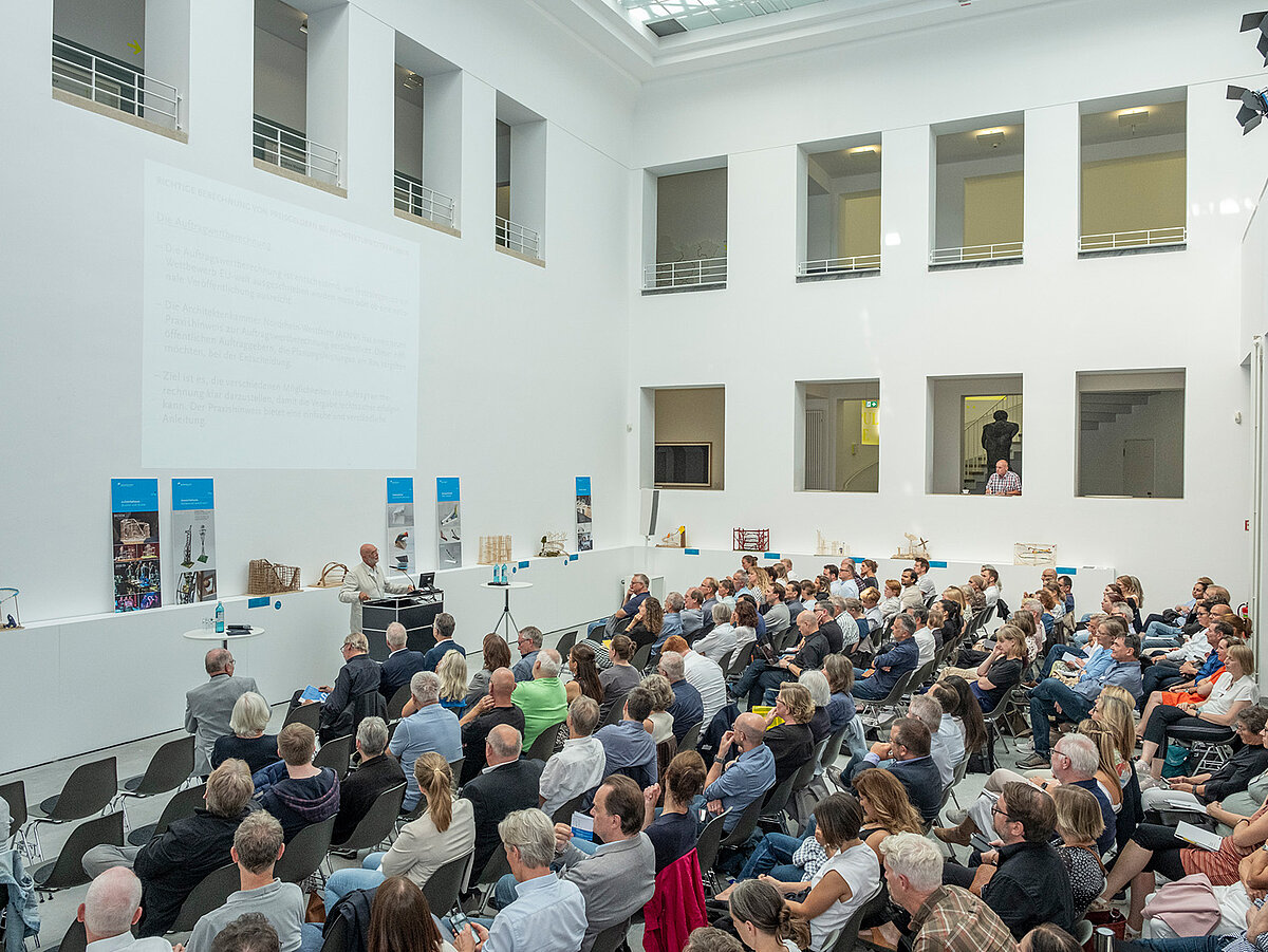 Ein heller Raum, viele Leute sitzen auf Stühlen im Publikum. An einem Pult steht eine Person und spricht zu dem Publikum. - Foto: Detlef Podehl