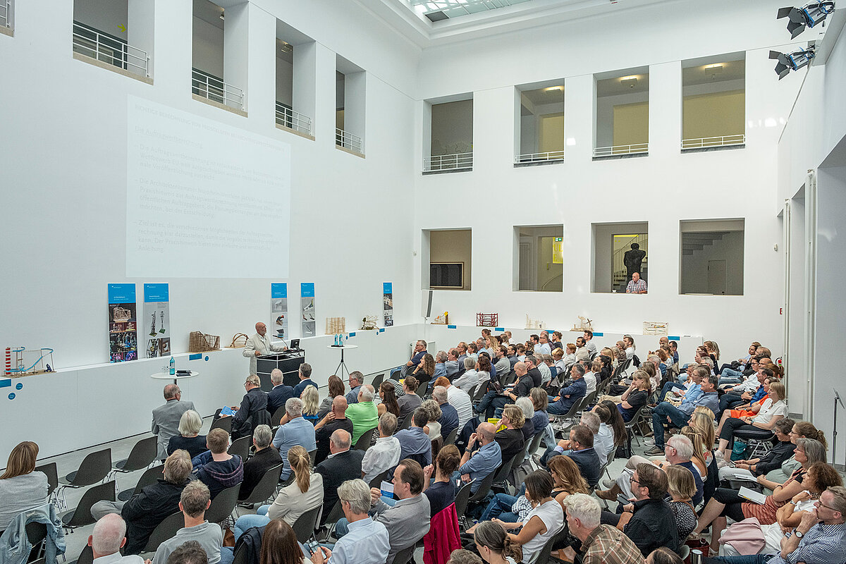 Ein heller Raum, viele Leute sitzen auf Stühlen im Publikum. An einem Pult steht eine Person und spricht zu dem Publikum. - Foto: Detlef Podehl