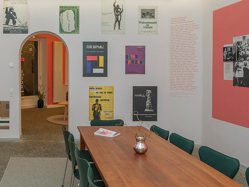 Ein Raum mit teilweise bunten rötlichen Wänden, teilweise weißen Wänden. An den Wänden hängen Bildtafeln und es sind Textschriftzüge befestigt. In der Mitte des Raumes steht ein Tisch mit Stühlen.