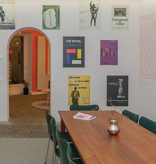 Ein Raum mit teilweise bunten rötlichen Wänden, teilweise weißen Wänden. An den Wänden hängen Bildtafeln und es sind Textschriftzüge befestigt. In der Mitte des Raumes steht ein Tisch mit Stühlen.