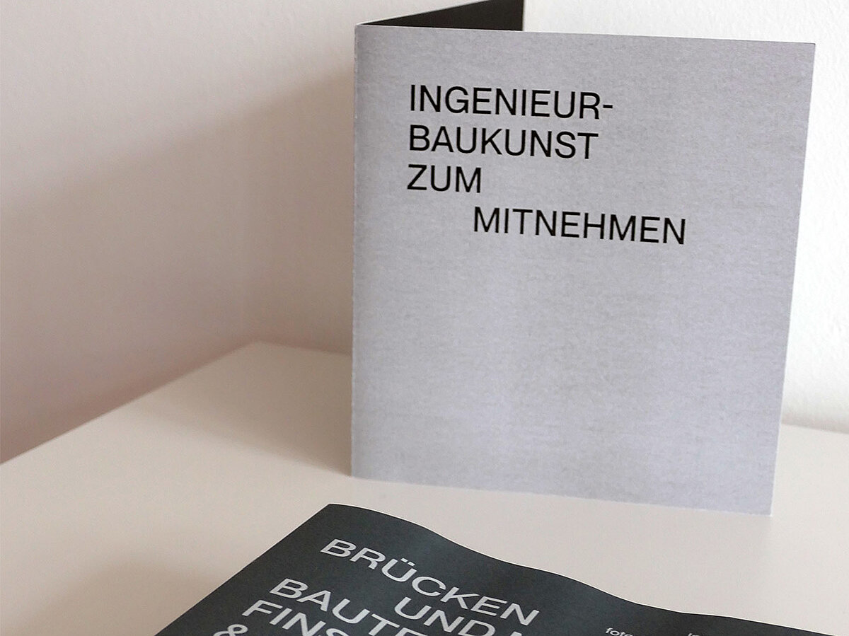 Eine Broschüre liegt auf einem Tisch, dahinter steht ein Schild mit Aufschrift.