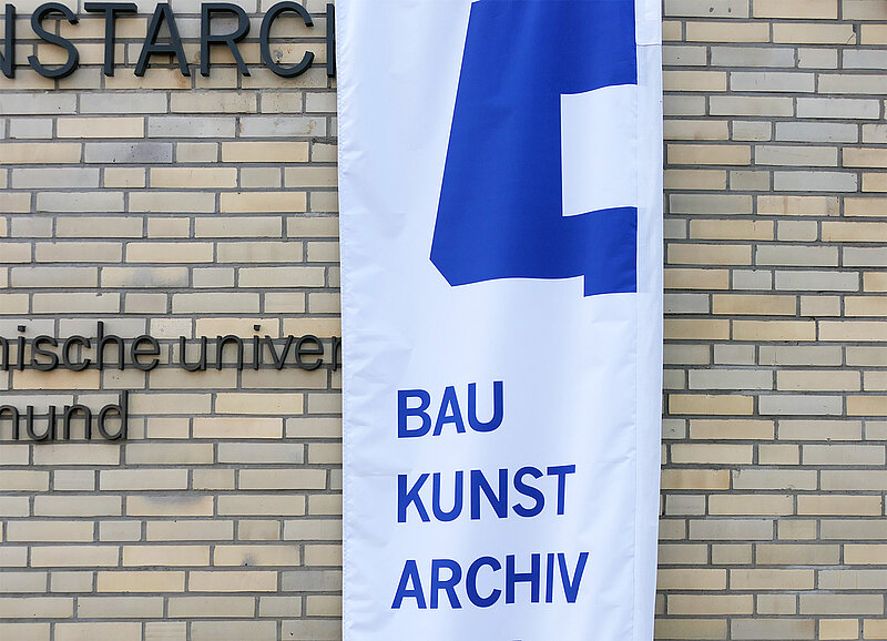 Blick auf eine helle Gebäudefassade, an der teilweise Textbuchstaben angebracht sind. Davor steht eine Flagge mit der Aufschrift "BAUKUNSTARCHIV"