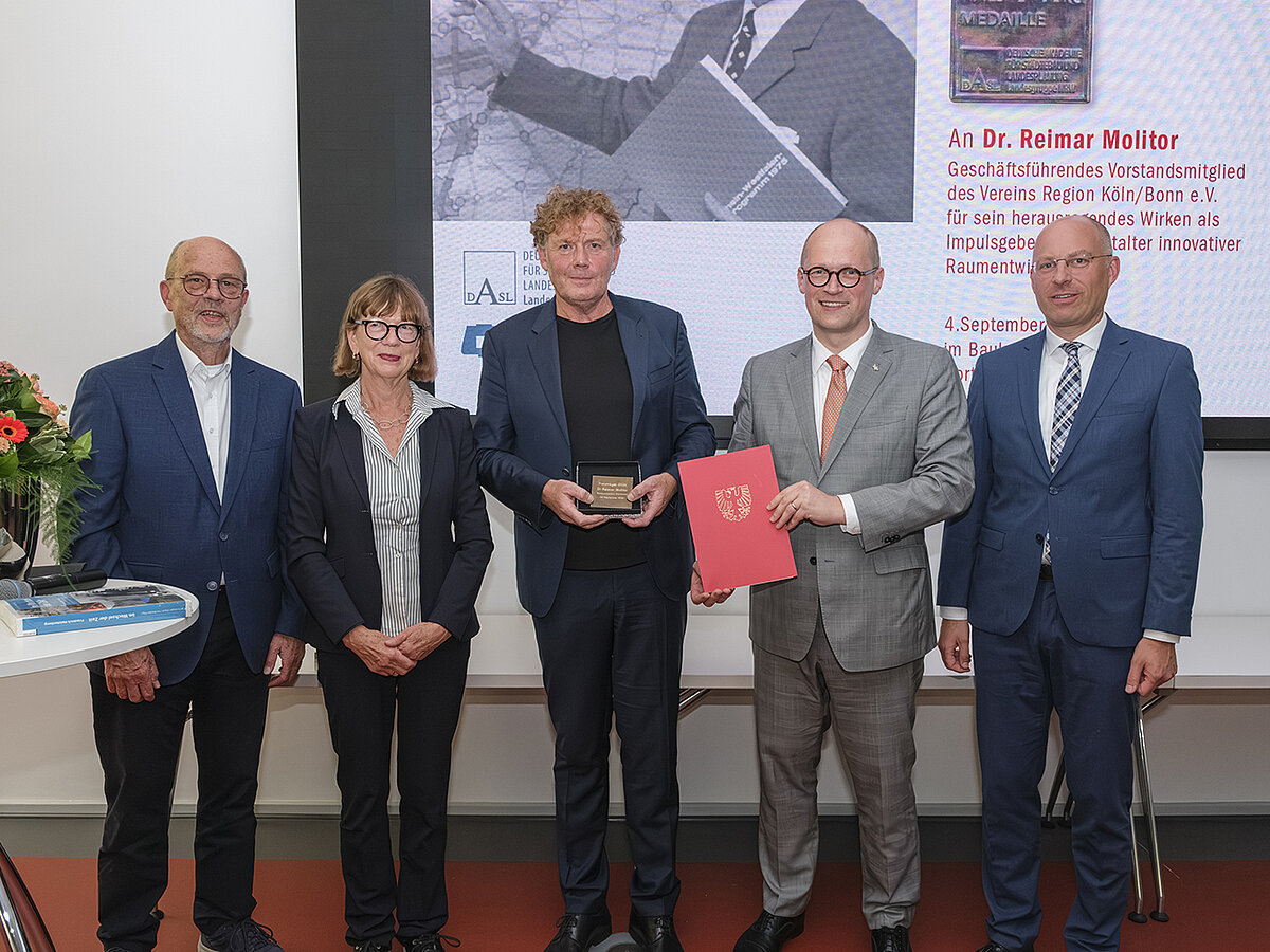 Personen stehen beisammen und lächeln in die Kamera. Im Hintergrund eine Leinwand mit Präsentation, neben den Personen ein Stehtisch mit Materialien drauf, unter anderem mit einem Blumenstrauß.
