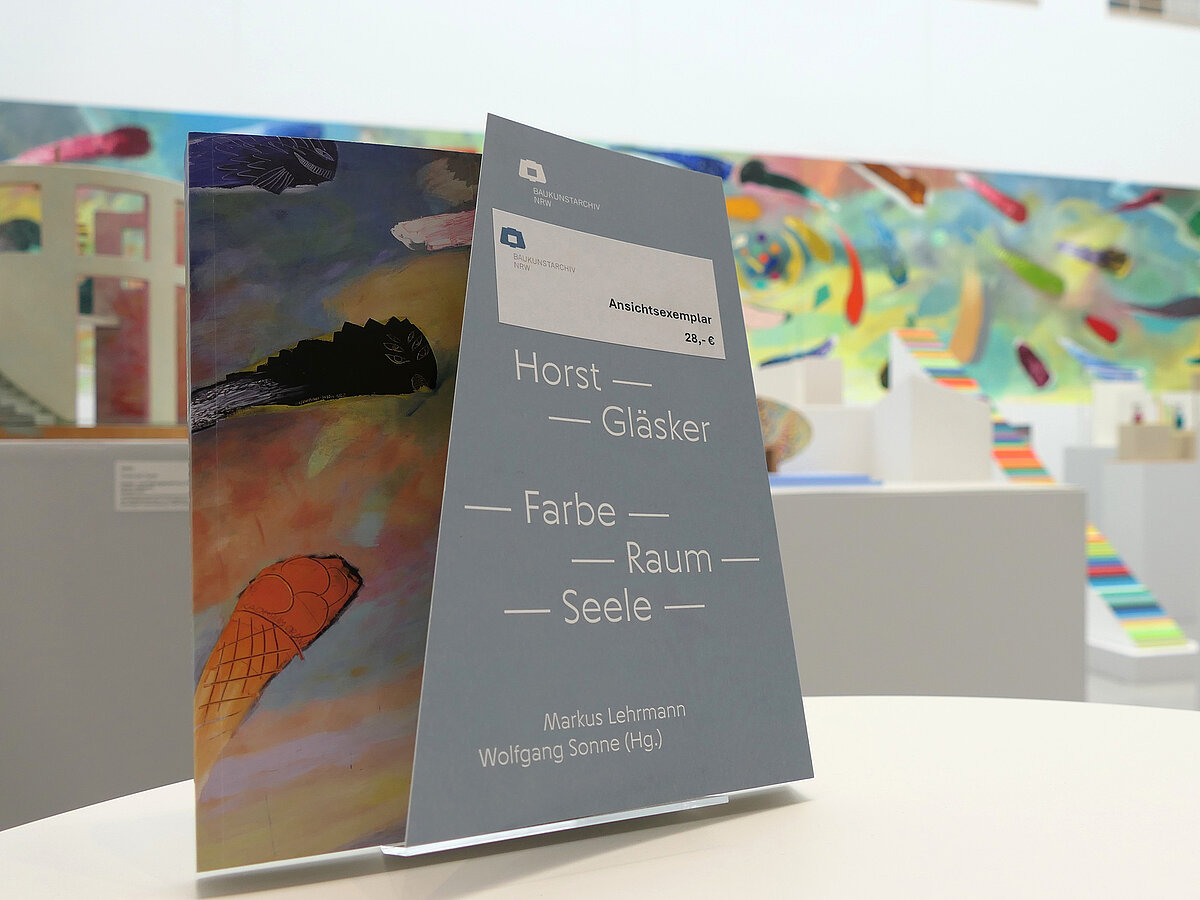 Ein Buch steht in einem Ausstellungsraum auf einem Tisch, dahinter sind Skulpturen und Gemälde platziert.
