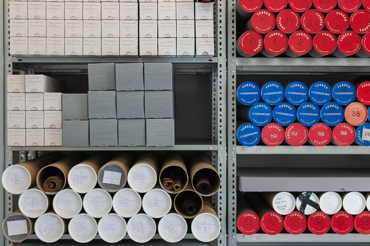 Regale, in denen Dokumente und Papiere platziert sind, teilweise in hellen, grauen und braunen Behältnissen, teilweise in blauen und roten runden Behältnissen. - Foto: Christine Kämmerer/Baukunstarchiv NRW 
