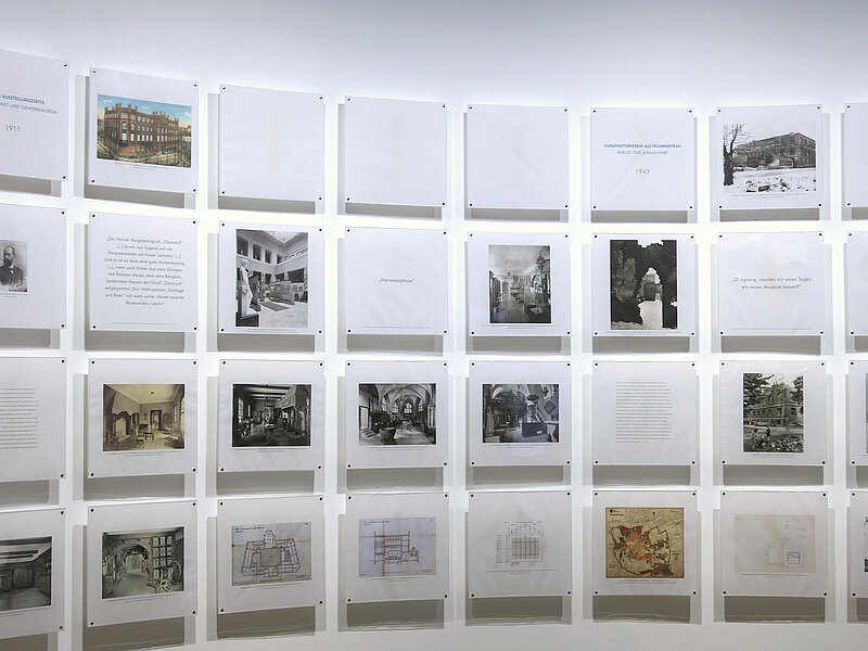 Eine weiße Wand, daran sind Foto- und Texttafeln befestigt.