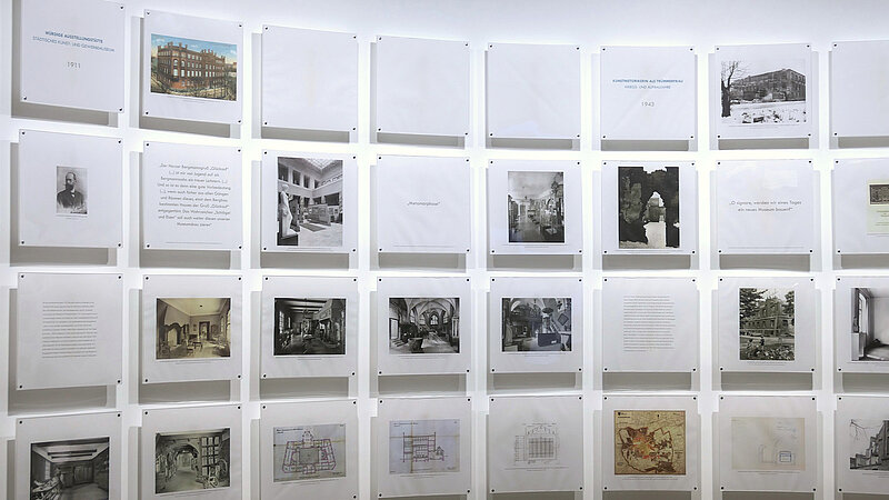 Eine weiße Wand, daran sind Foto- und Texttafeln befestigt.
