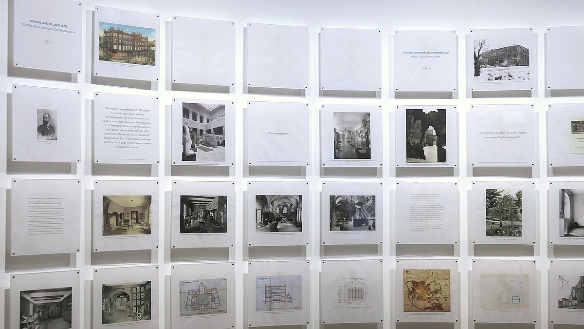 Eine weiße Wand, daran sind Foto- und Texttafeln befestigt.
