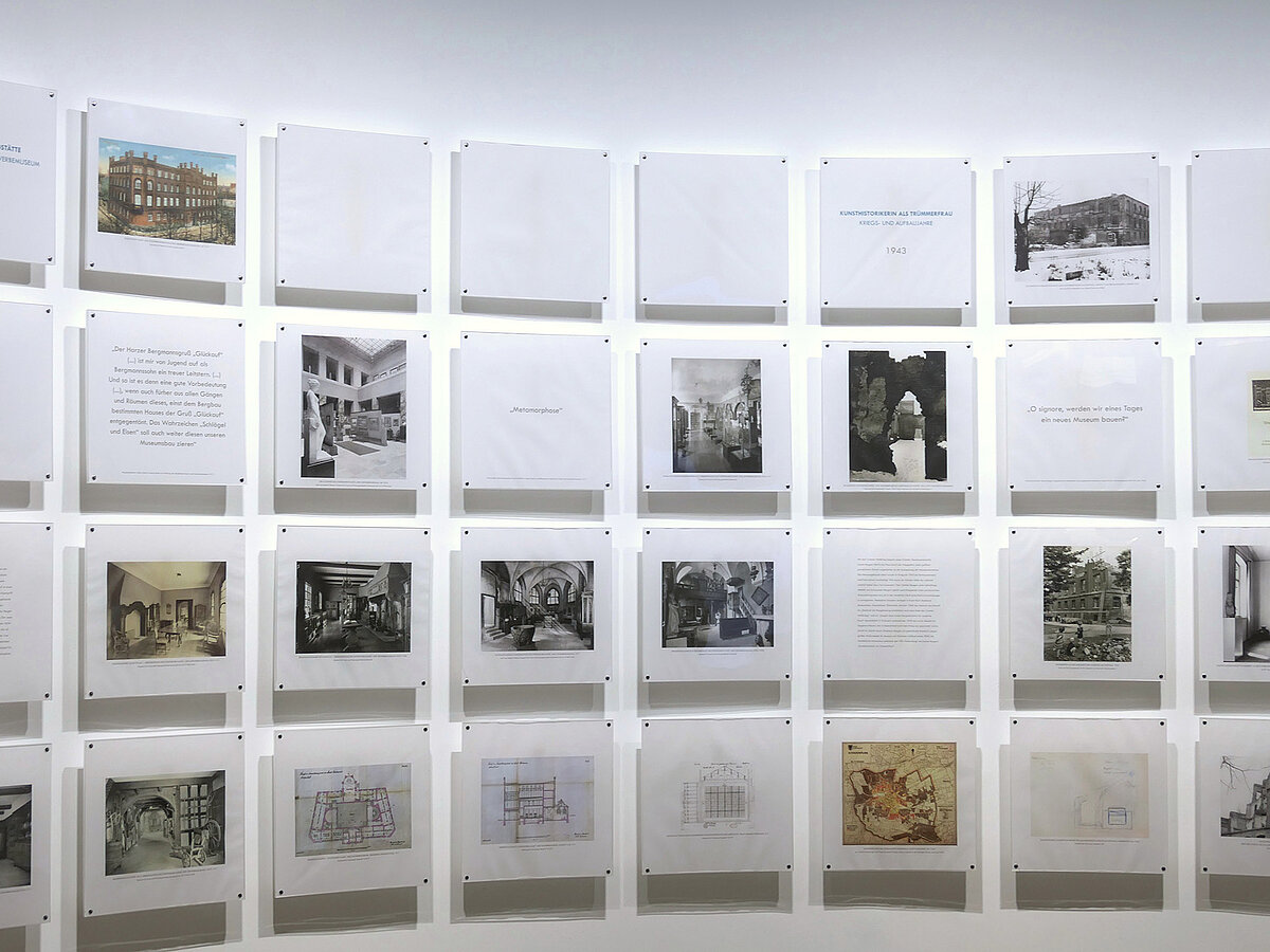 Eine weiße Wand, daran sind Foto- und Texttafeln befestigt.