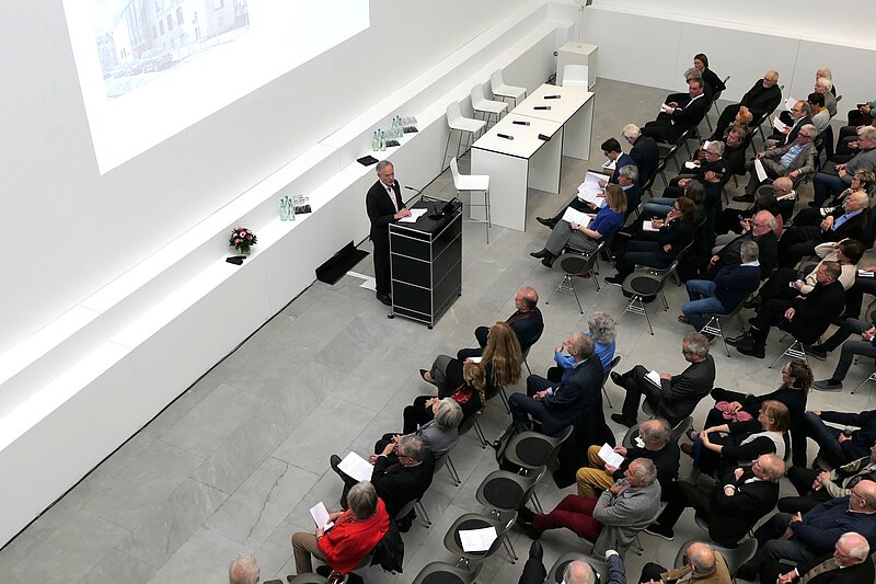 Ein heller Raum, viele Leute sitzen auf Stühlen im Publikum. An einem Pult steht eine Person und spricht zu dem Publikum. Hinter dem Pult wird eine Präsentation per Beamer auf der Wand präsentiert. Das Bild wurde aus der Vogelperspektive aufgenommen. 