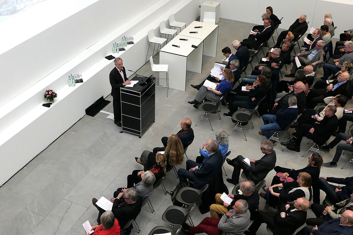 Ein heller Raum, viele Leute sitzen auf Stühlen im Publikum. An einem Pult steht eine Person und spricht zu dem Publikum. Hinter dem Pult wird eine Präsentation per Beamer auf der Wand präsentiert. Das Bild wurde aus der Vogelperspektive aufgenommen. 