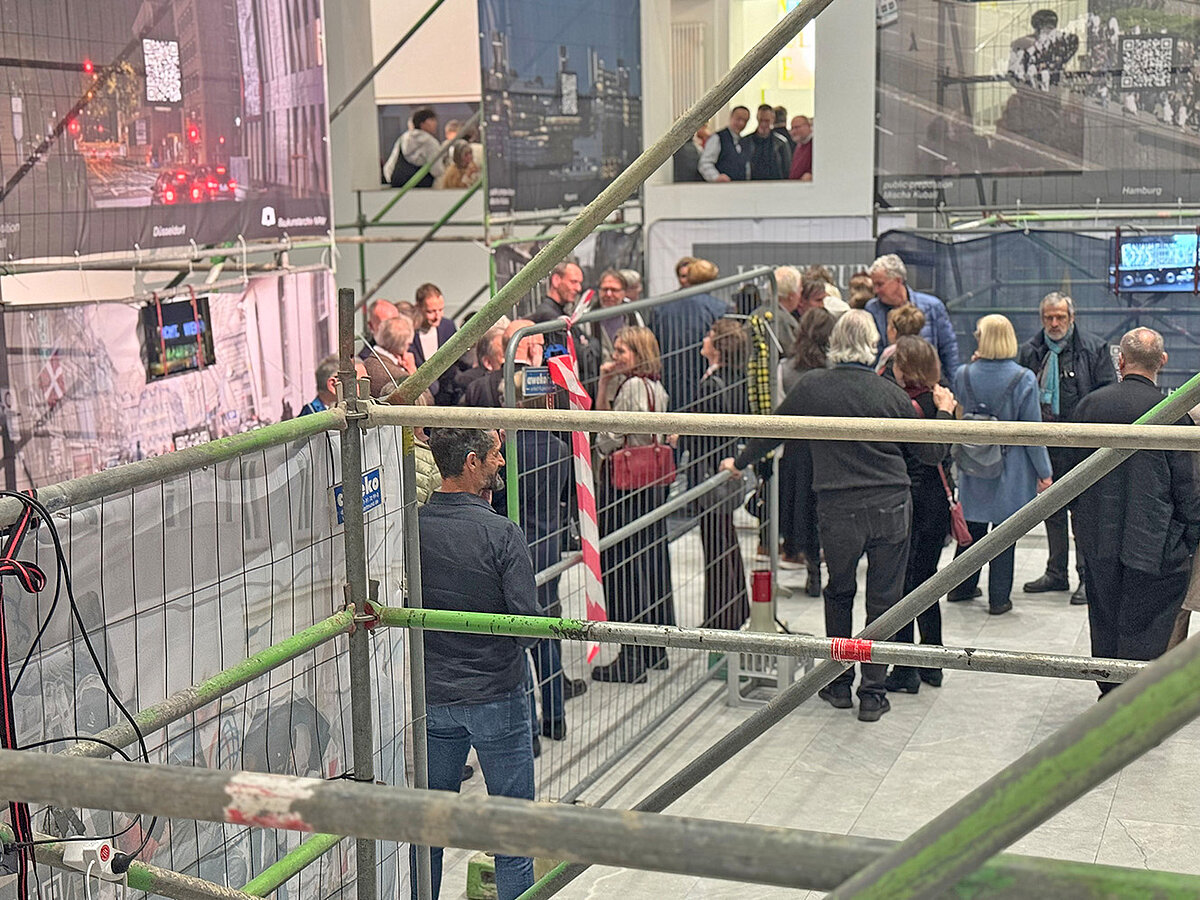 Personen stehen in einem geschlossenen Raum mit Bauzäunen, die teilweise mit Transparenten bespannt sind.
